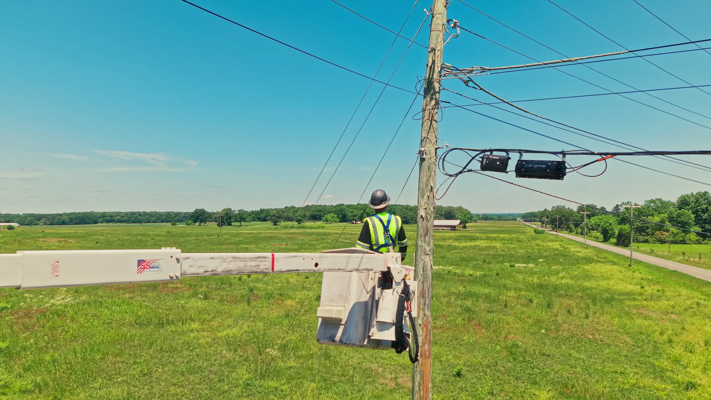NextGen Fiber lineman in the bucket at a pole line with aerial splice enclosures, rural road in the background